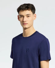 A person with short dark hair wears the MENS NICHOLSON PB CHEST GRAPHIC TEE from Psycho Bunny, a navy blue Pima cotton T-shirt with embroidered chest details, set against a plain light background.