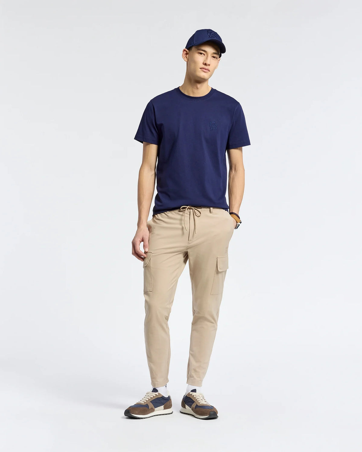 A person in a navy Psycho Bunny MENS NICHOLSON PB CHEST GRAPHIC TEE, navy cap, beige regular-fit cargo jogger pants, and sneakers stands against a white background, posed relaxedly with hands in pockets, looking at the camera.