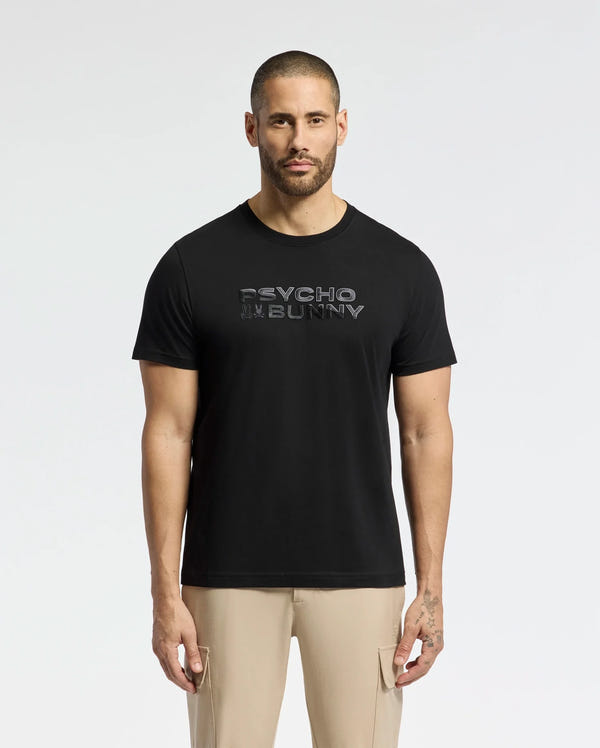 A man with a buzz cut wears a black MENS BERNI EMBROIDERED WORDMARK GRAPHIC TEE from Psycho Bunny, crafted from plush Pima cotton. "Psycho Bunny" is embroidered on the logo. He pairs it with beige pants and stands against a plain white background.
