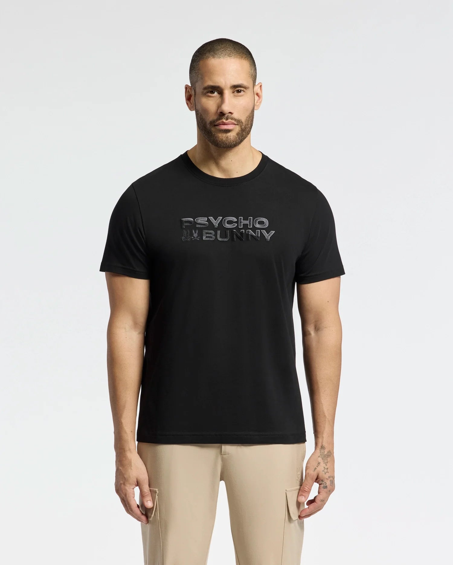 A man with a buzz cut wears a black MENS BERNI EMBROIDERED WORDMARK GRAPHIC TEE from Psycho Bunny, crafted from plush Pima cotton. "Psycho Bunny" is embroidered on the logo. He pairs it with beige pants and stands against a plain white background.