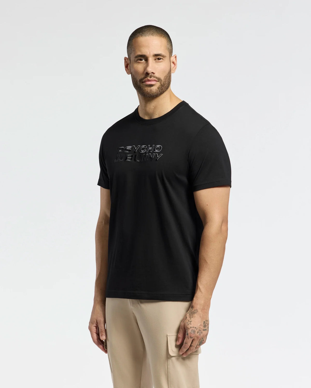 A man with a shaved head and beard faces forward in a black Psycho Bunny "MENS BERNI EMBROIDERED WORDMARK GRAPHIC TEE - B6U417F200" made of Pima cotton. He pairs it with beige pants, showing his tattooed left arm against a plain white background.