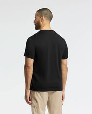 A man with short hair, viewed from the back, wears a black Psycho Bunny Men's Berni Embroidered Wordmark Graphic Tee made from soft Pima cotton and beige pants. He stands against a plain white background, showcasing the tee's subtle elegance.