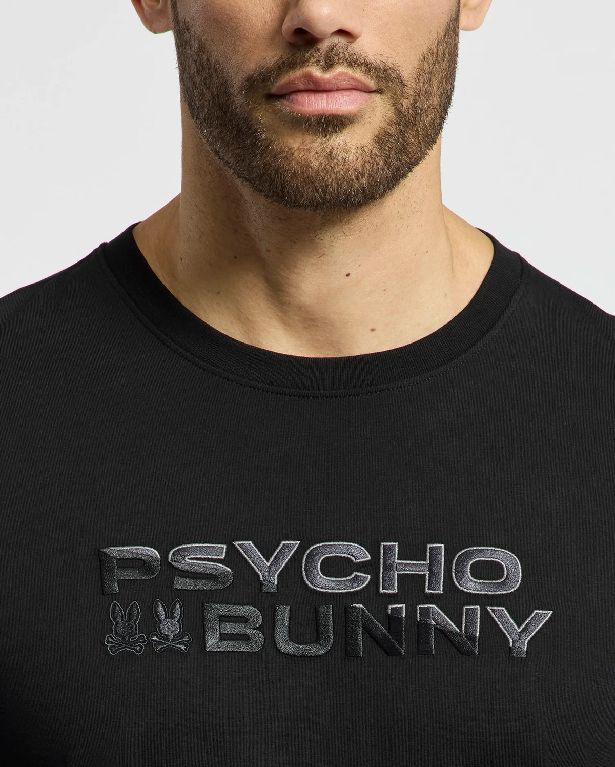 A person wearing the Psycho Bunny Men's Berni Embroidered Wordmark Graphic Tee in black Pima cotton, featuring raised silver "PSYCHO BUNNY" text and small embroidered bunny logos, their face partially visible with a trimmed beard.