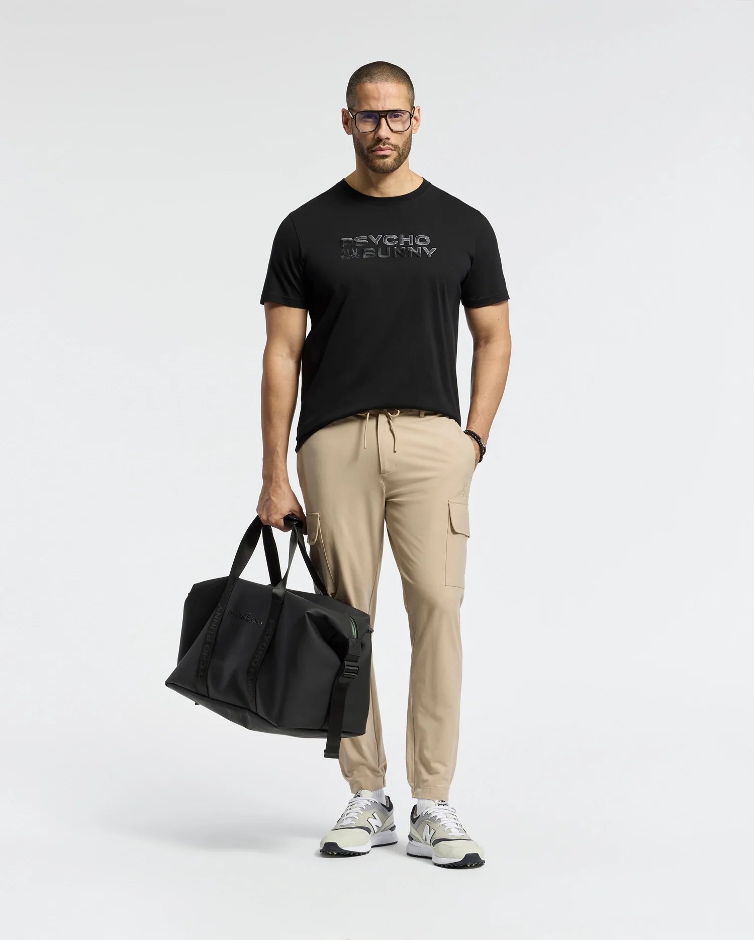 A man with glasses confidently stands against a light background, wearing a Psycho Bunny MENS BERNI EMBROIDERED WORDMARK GRAPHIC TEE, beige cargo pants, and white sneakers while holding a large black duffel bag in his left hand.