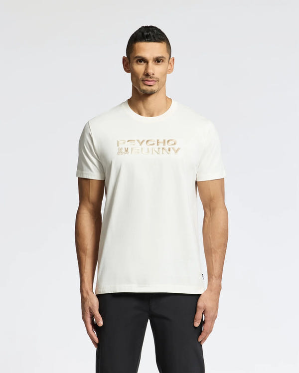 A man with short dark hair stands against a white backdrop, wearing black pants and the Psycho Bunny Men's Berni Embroidered Wordmark Graphic Tee (B6U417F200), a white Pima cotton T-shirt featuring "Psycho Bunny" in gold lettering across the chest with an embroidered logo.