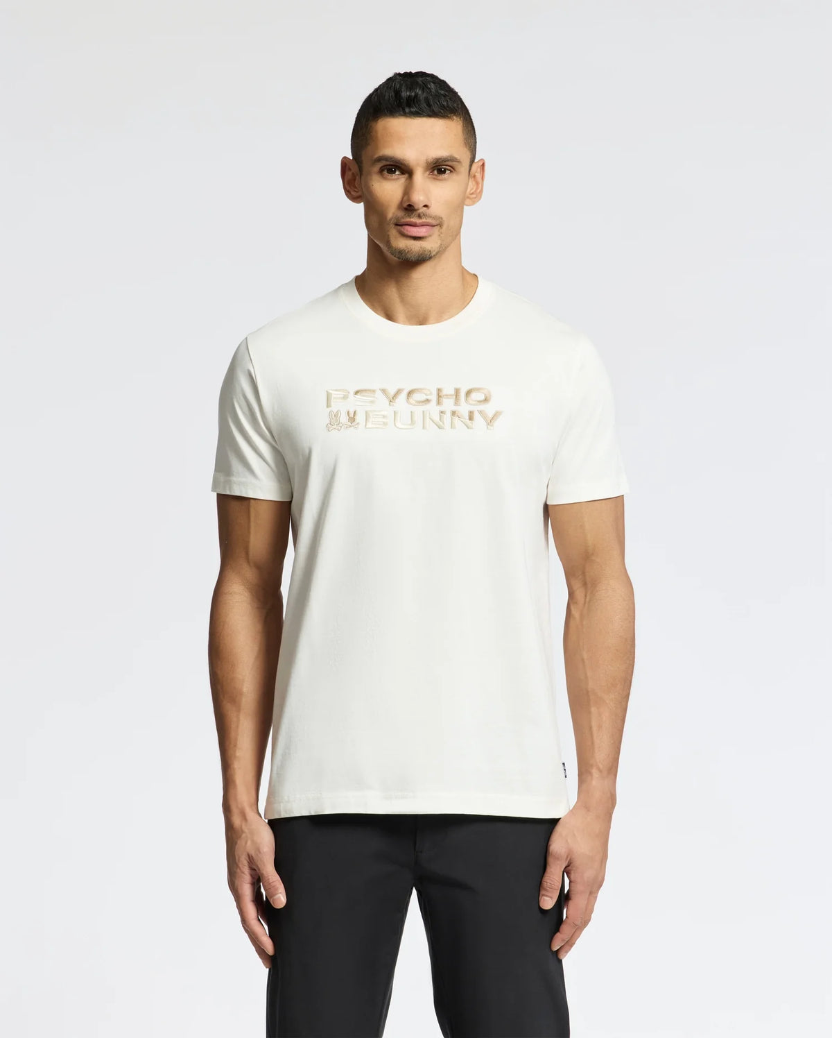 A man with short dark hair stands against a white backdrop, wearing black pants and the Psycho Bunny Men's Berni Embroidered Wordmark Graphic Tee (B6U417F200), a white Pima cotton T-shirt featuring "Psycho Bunny" in gold lettering across the chest with an embroidered logo.