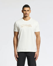 A man with short dark hair stands against a white backdrop, wearing black pants and the Psycho Bunny Men's Berni Embroidered Wordmark Graphic Tee (B6U417F200), a white Pima cotton T-shirt featuring "Psycho Bunny" in gold lettering across the chest with an embroidered logo.