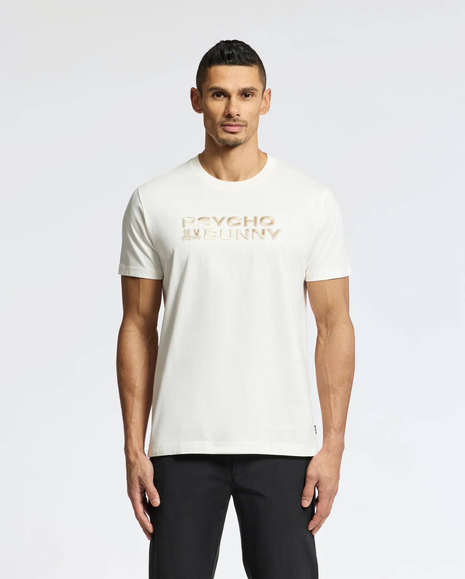 A man with short dark hair stands against a white backdrop, wearing black pants and the Psycho Bunny Men's Berni Embroidered Wordmark Graphic Tee (B6U417F200), a white Pima cotton T-shirt featuring "Psycho Bunny" in gold lettering across the chest with an embroidered logo.