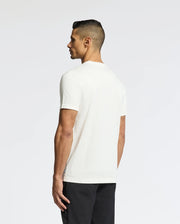 A man with short dark hair, facing away from the camera, wears a Psycho Bunny MENS BERNI EMBROIDERED WORDMARK GRAPHIC TEE in plain white with an embroidered logo, paired with black pants against a light gray background.