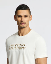 A man with short dark hair and facial hair wears the Psycho Bunny Men's Berni Embroidered Wordmark Graphic Tee, featuring metallic "PSYCHO BUNNY" text and an embroidered logo, as he stands against a plain white background.