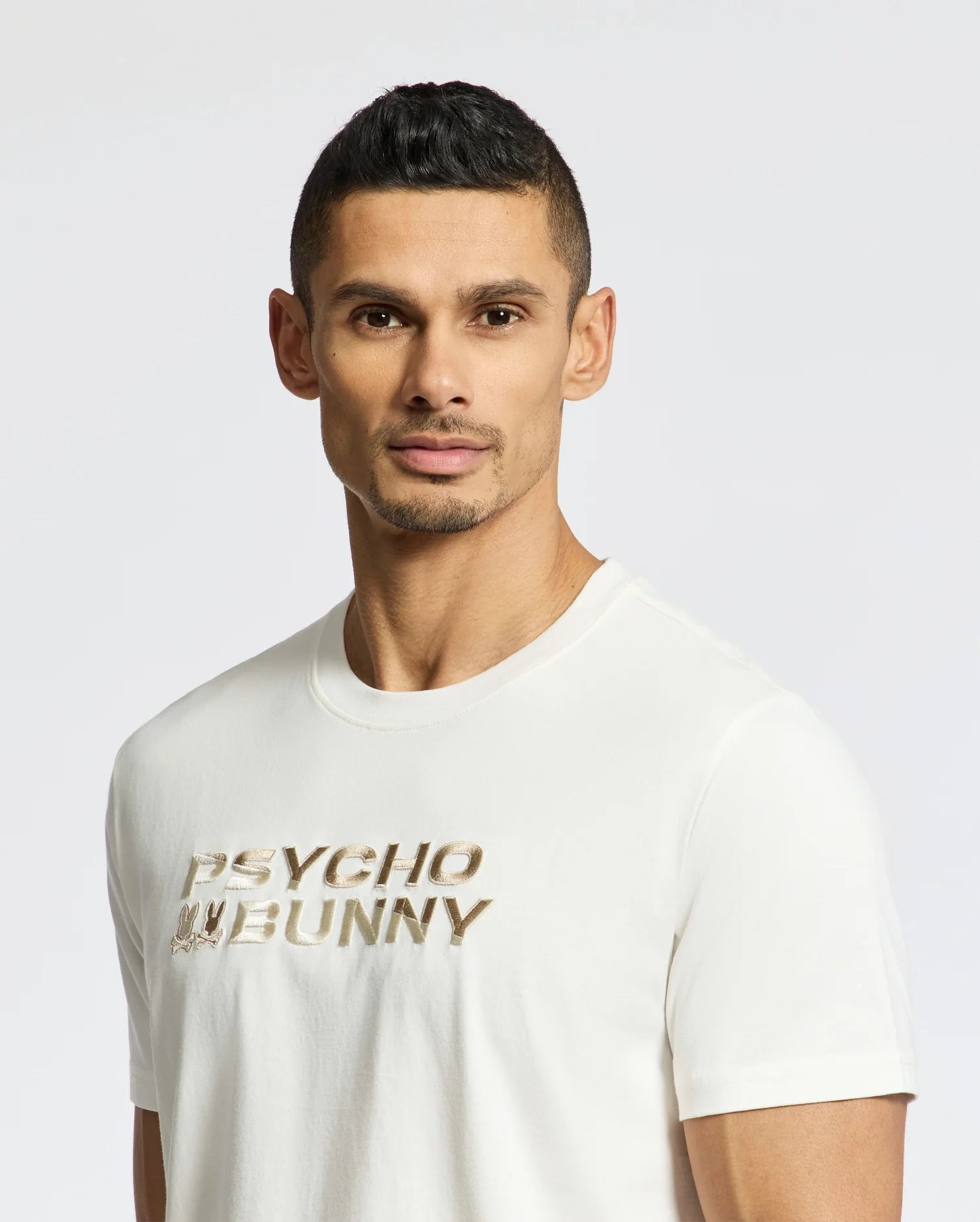 A man with short dark hair and facial hair wears the Psycho Bunny Men's Berni Embroidered Wordmark Graphic Tee, featuring metallic "PSYCHO BUNNY" text and an embroidered logo, as he stands against a plain white background.