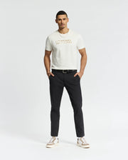 A man stands against a plain background wearing a white Psycho Bunny Mens Berni Embroidered Wordmark Graphic Tee, crafted from Pima cotton, paired with black pants and white sneakers. His hands rest in his pockets as he looks directly at the camera.