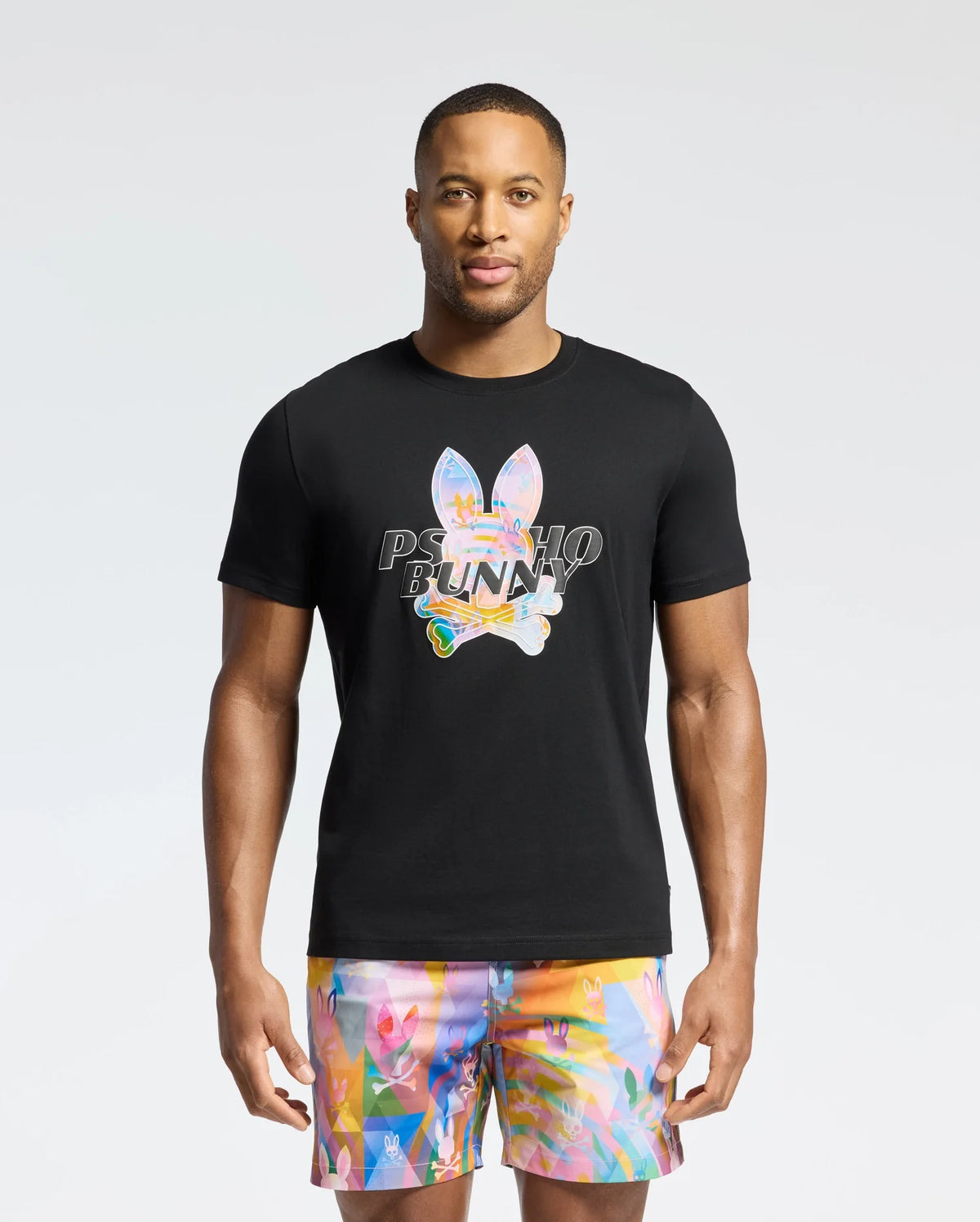 A person dons the Psycho Bunny MENS MILOS GRAPHIC TEE - B6U456F200 in black, featuring a bright bunny graphic and "Psycho Bunny" text, paired with multicolored abstract shorts against a plain white backdrop.