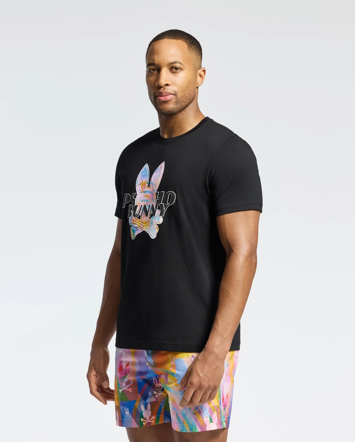 A person in a black Psycho Bunny MENS MILOS GRAPHIC TEE - B6U456F200, made of luxurious Peruvian Pima cotton with a colorful bunny graphic, stands facing slightly left, wearing vibrant multicolored shorts against a plain white background.