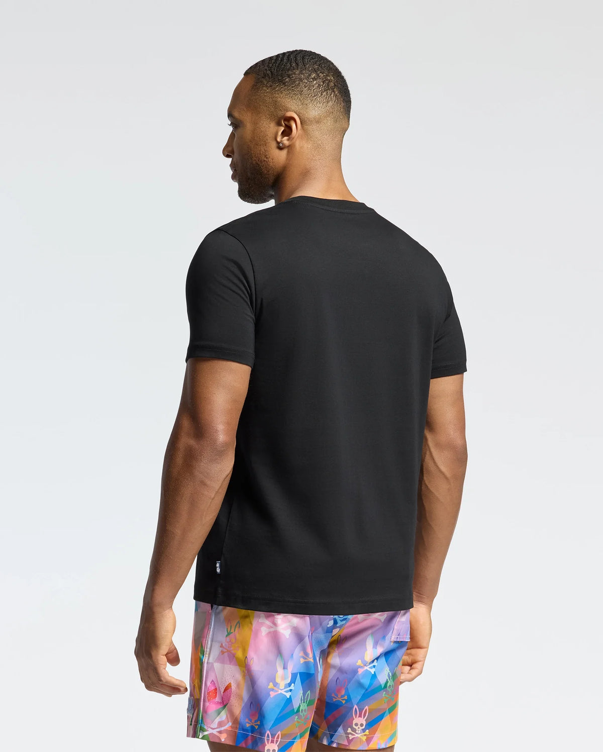 A man wearing the Psycho Bunny MENS MILOS GRAPHIC TEE in black and vibrant floral-patterned shorts stands with his back to the camera. The bright, uncluttered background highlights his casual, futuristic style.