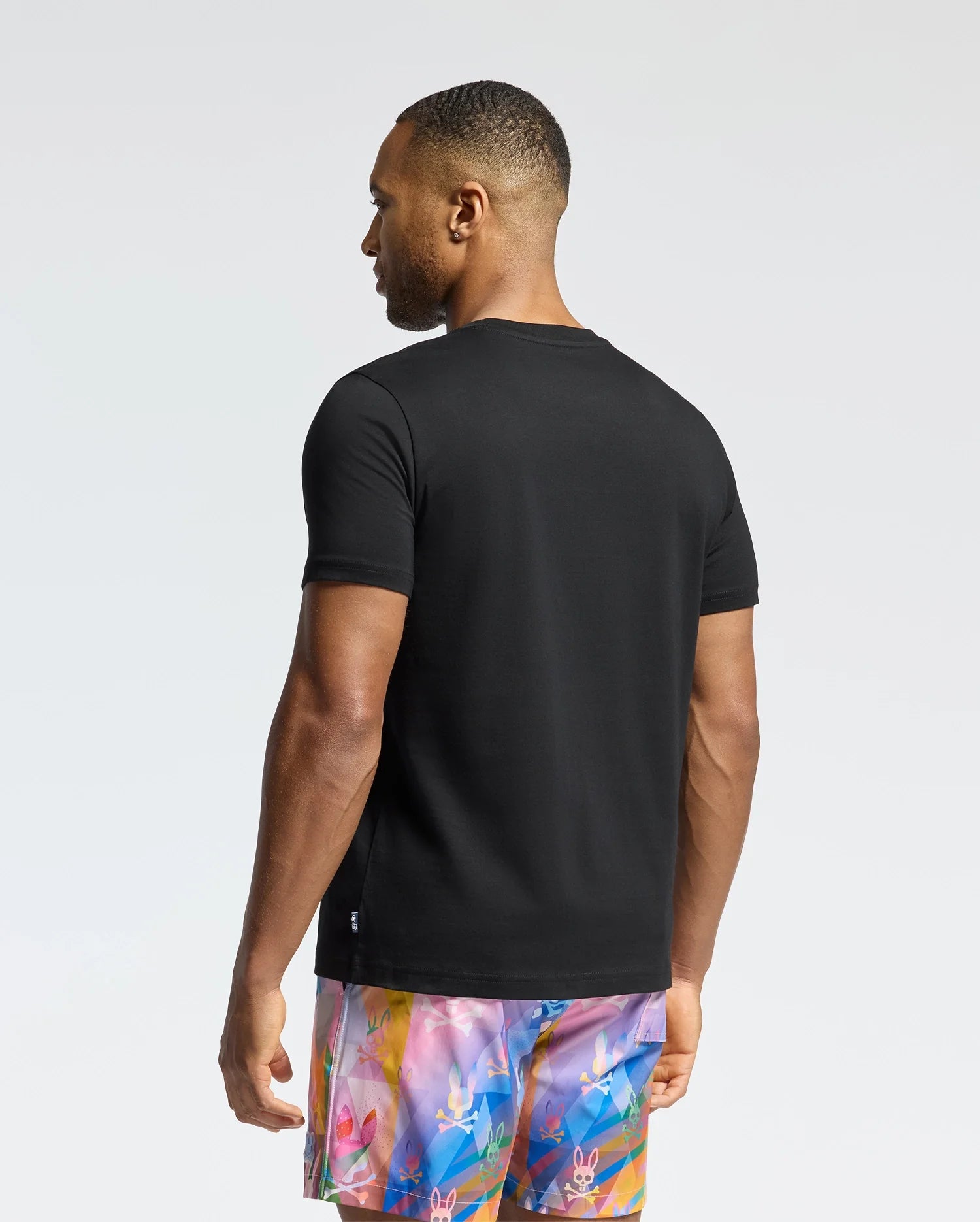A man wearing the Psycho Bunny MENS MILOS GRAPHIC TEE in black and vibrant floral-patterned shorts stands with his back to the camera. The bright, uncluttered background highlights his casual, futuristic style.