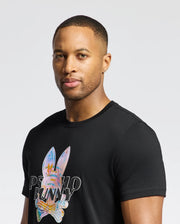 A person in a black Psycho Bunny MENS MILOS GRAPHIC TEE - B6U456F200, made from luxurious Peruvian Pima cotton, with a colorful bunny graphic and "PSYCHO BUNNY" text, is set against a plain white background.