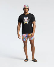 A man confidently stands against a gray background wearing the Psycho Bunny MENS MILOS GRAPHIC TEE - B6U456F200 in black. His colorful shorts and matching bucket hat add flair, while black slide sandals complete his look, effortlessly merging casual style with playful charm.