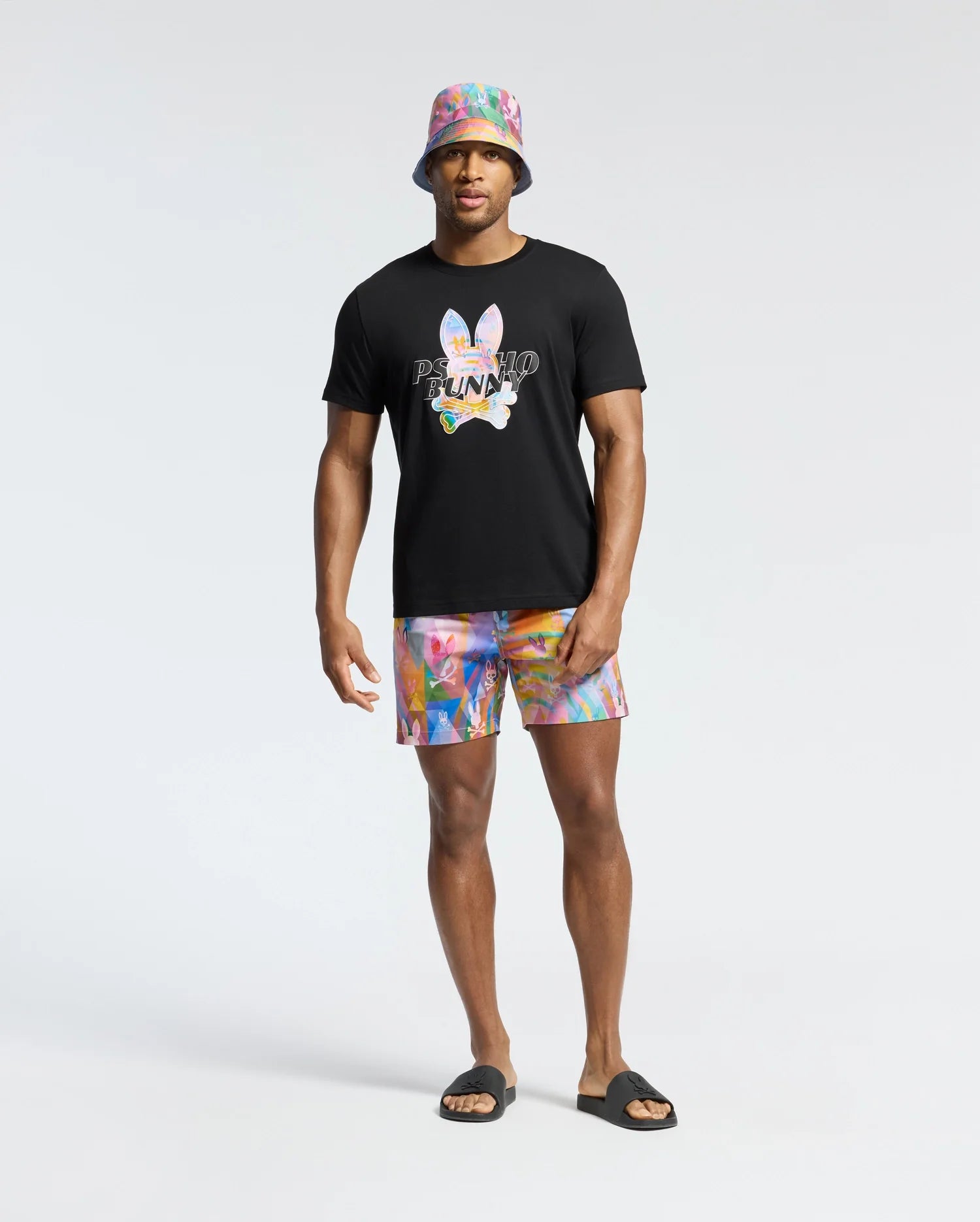 A man confidently stands against a gray background wearing the Psycho Bunny MENS MILOS GRAPHIC TEE - B6U456F200 in black. His colorful shorts and matching bucket hat add flair, while black slide sandals complete his look, effortlessly merging casual style with playful charm.