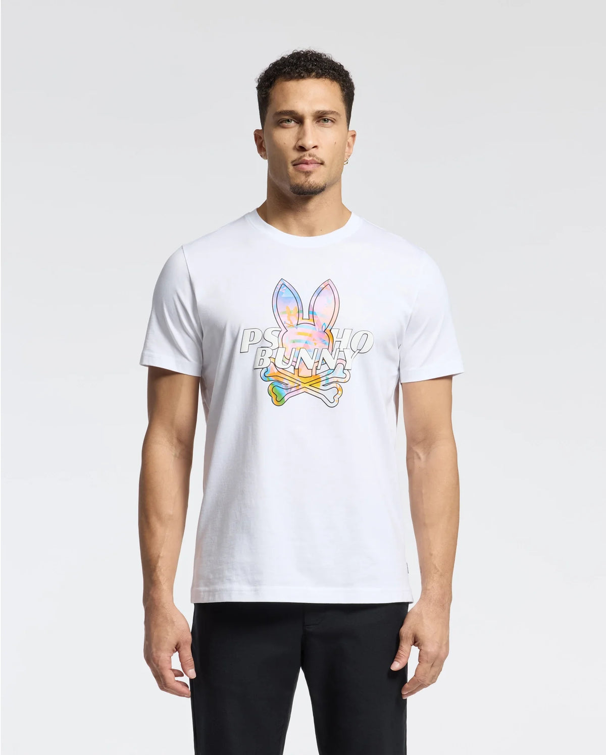 A man in a white Psycho Bunny MENS MILOS GRAPHIC TEE, made from Peruvian Pima cotton with a space-age graphic and bunny logo, stands against a plain background. His short curly hair complements his direct gaze at the camera, paired with sleek black pants.