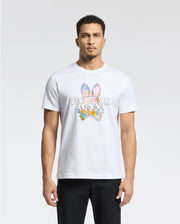 A man in a white Psycho Bunny MENS MILOS GRAPHIC TEE, made from Peruvian Pima cotton with a space-age graphic and bunny logo, stands against a plain background. His short curly hair complements his direct gaze at the camera, paired with sleek black pants.