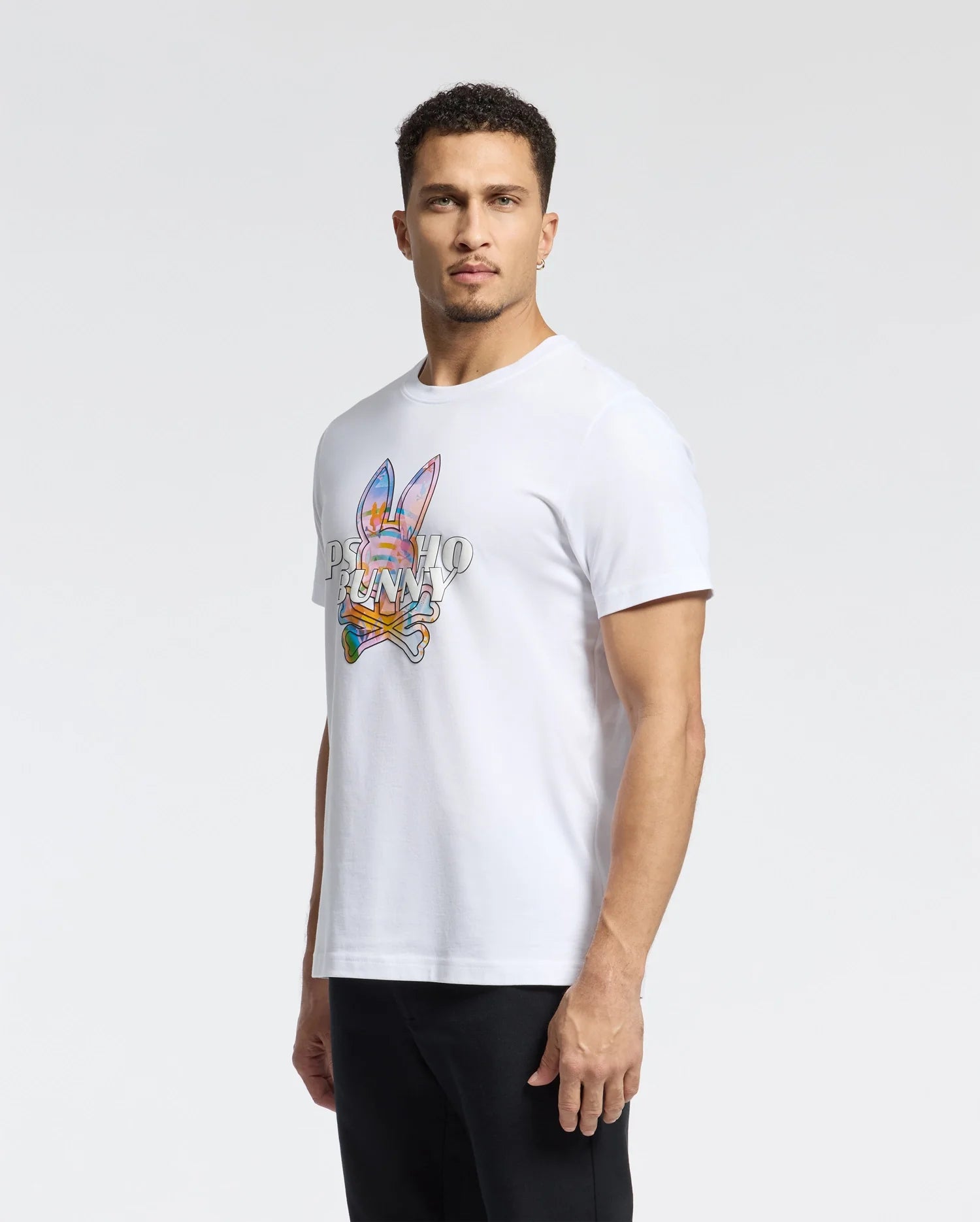 A man in a Psycho Bunny MENS MILOS GRAPHIC TEE - B6U456F200, featuring a space-age design, stands against a plain background. Crafted from luxurious Peruvian Pima cotton, it pairs perfectly with his black pants. His short hair complements the modern vibe of the look.