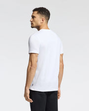 A man with short curly hair, wearing a Psycho Bunny MENS MILOS GRAPHIC TEE - B6U456F200 and black pants, stands facing left and glances over his shoulder against a plain background.