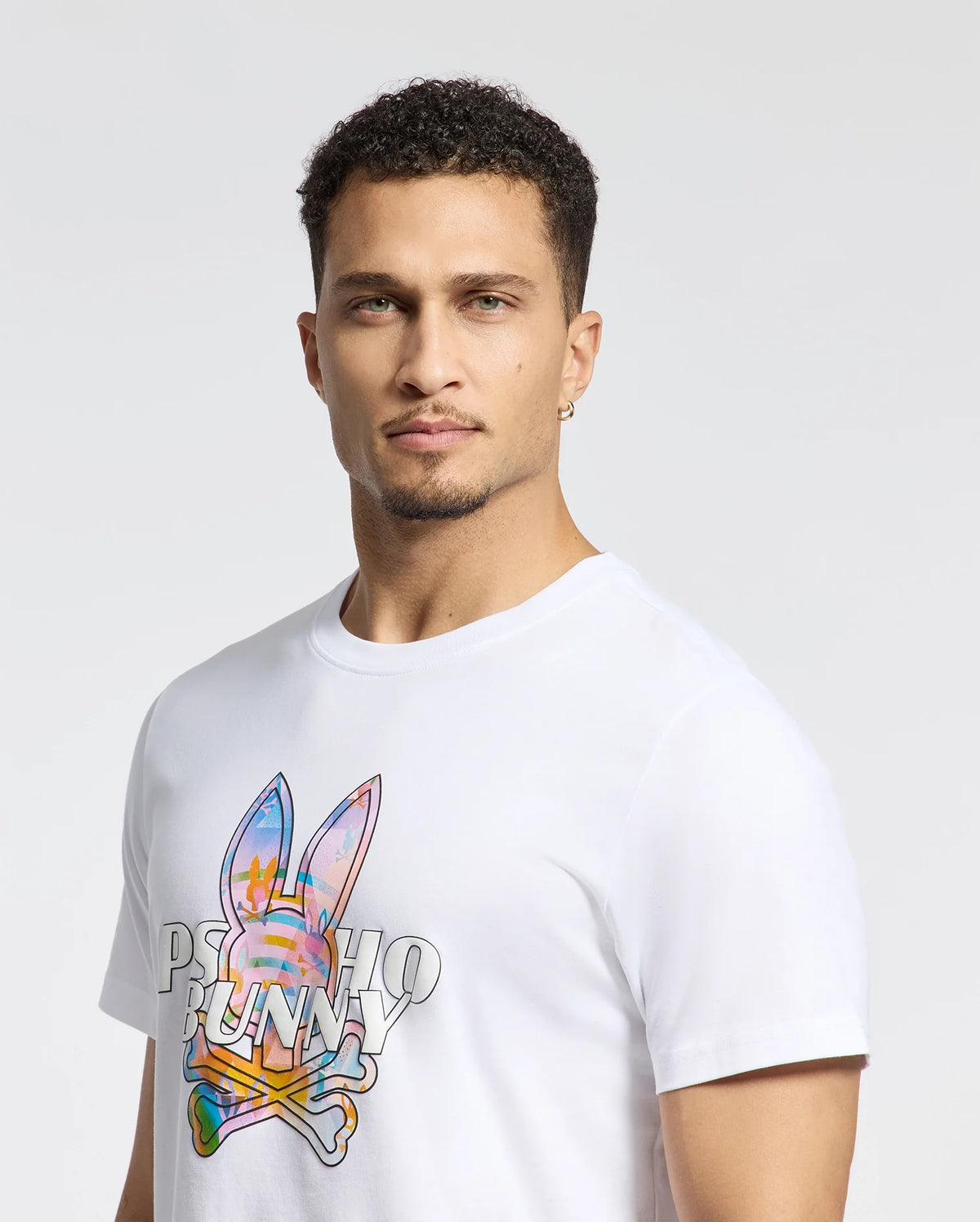 A man with short curly hair wears Psycho Bunny's MENS MILOS GRAPHIC TEE (B6U456F200), a white shirt made of Peruvian Pima cotton featuring a colorful space-age bunny graphic and "PSYCHO BUNNY" text. He has a neutral expression, a small hoop earring in his left ear, against a light background.