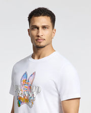 A man with short curly hair wears Psycho Bunny's MENS MILOS GRAPHIC TEE (B6U456F200), a white shirt made of Peruvian Pima cotton featuring a colorful space-age bunny graphic and "PSYCHO BUNNY" text. He has a neutral expression, a small hoop earring in his left ear, against a light background.