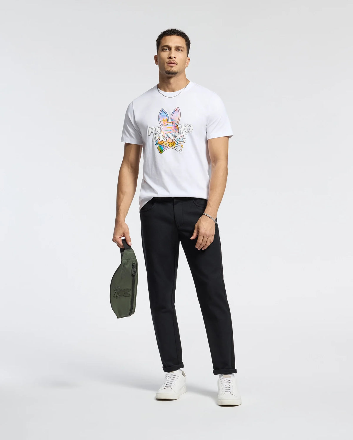A man stands against a plain background wearing the Psycho Bunny MENS MILOS GRAPHIC TEE - B6U456F200, black pants, and white sneakers. He holds a small green bag in his left hand, effortlessly blending style and comfort.
