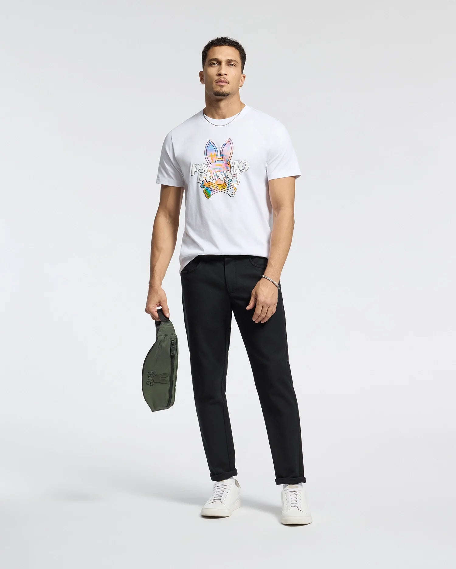 A man stands against a plain background wearing the Psycho Bunny MENS MILOS GRAPHIC TEE - B6U456F200, black pants, and white sneakers. He holds a small green bag in his left hand, effortlessly blending style and comfort.