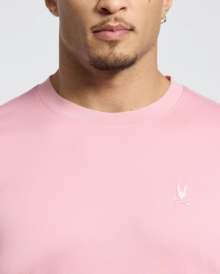 A person is seen from the shoulders up wearing the Psycho Bunny MEN'S BRYCE PERFORMANCE TEE (B6U490F200) in pink, made of a Modal-Pima cotton blend from Peru. The chest features an embroidered logo with a stylized rabbit head and crossed bones. They have short hair, stubble, and an earring.