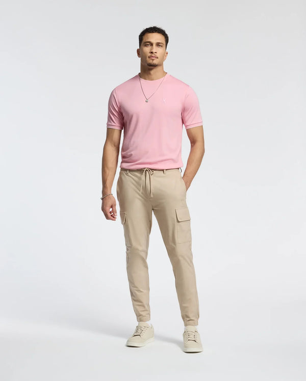 A man stands confidently in a Psycho Bunny MENS BRYCE PERFORMANCE TEE - B6U490F200, made from a Modal-Pima cotton blend, paired with tan cargo joggers, beige sneakers, and a silver necklace. The plain light background highlights his casual yet stylish look.