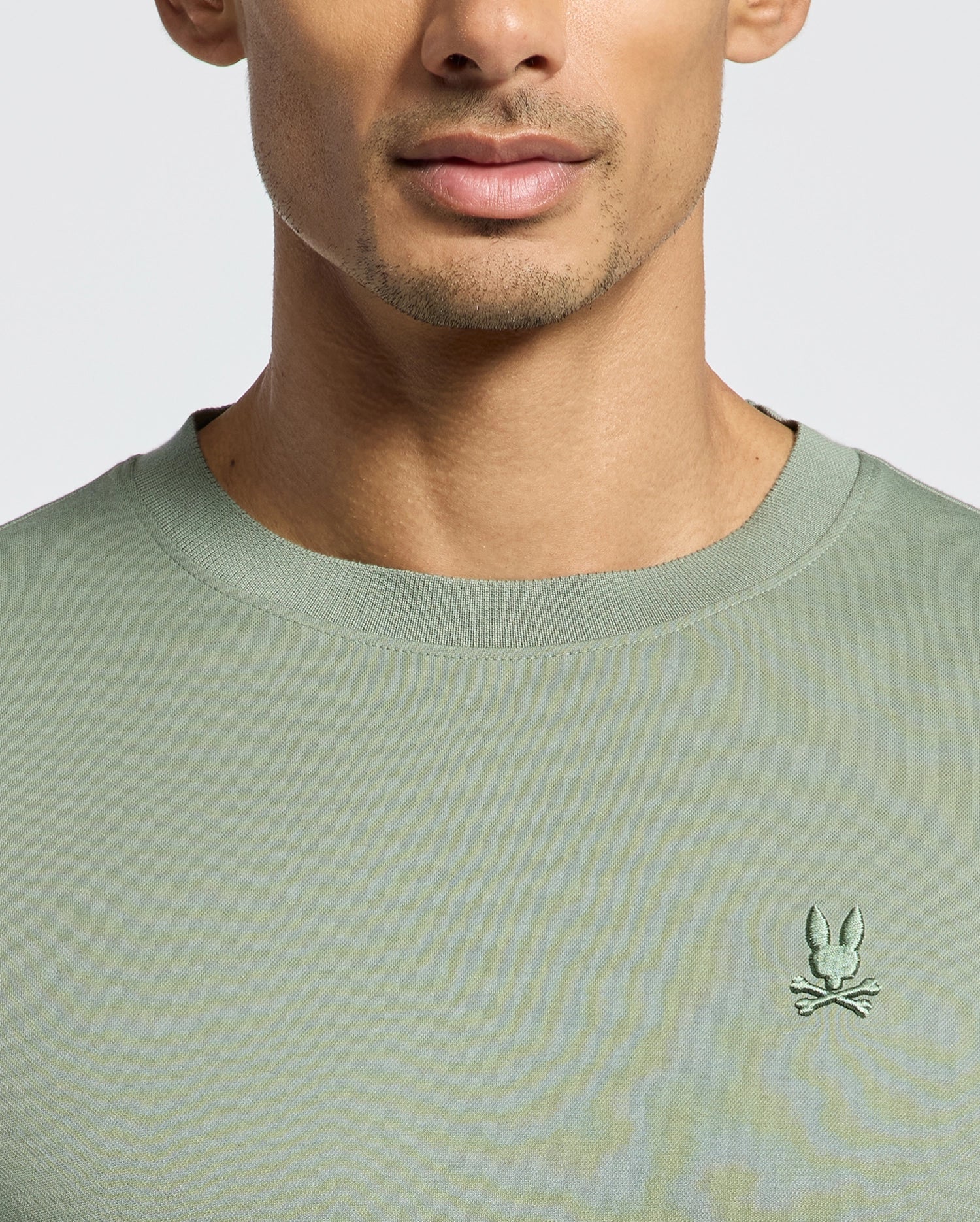 Someone wearing the green Psycho Bunny MENS BRYCE PERFORMANCE TEE, made from soft Peruvian Pima cotton, featuring a small embroidered bunny and crossed bones emblem on the chest, with their face partially visible against a plain background.