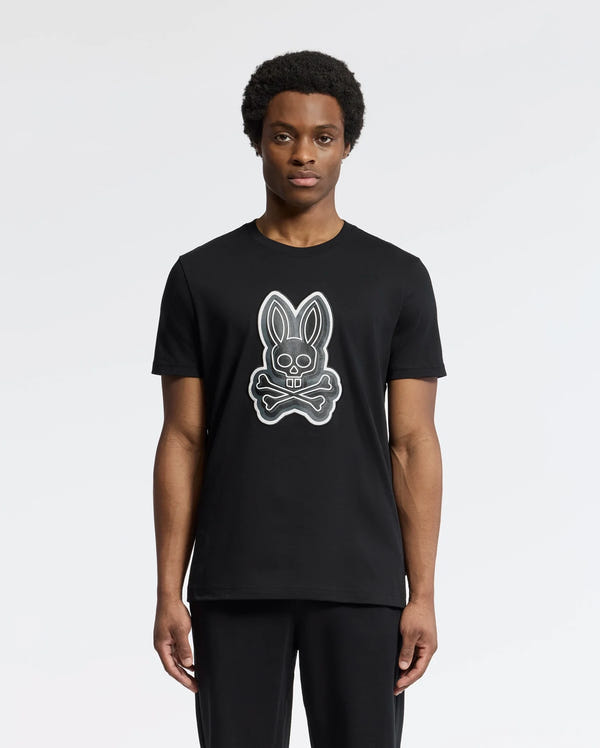 A person wearing the Psycho Bunny MENS SYDNEY GRAPHIC TEE - B6U508F200, featuring a cartoonish bunny and crossbones graphic print on black Pima cotton, stands against a plain white background. With short curly hair and a neutral expression, they showcase the casual charm of its regular fit.