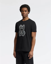 A person is wearing the Psycho Bunny MENS SYDNEY GRAPHIC TEE - B6U508F200, a black T-shirt made from soft Pima cotton. It features a graphic print of a bunny and crossbones on the front in regular fit, set against a plain white background.