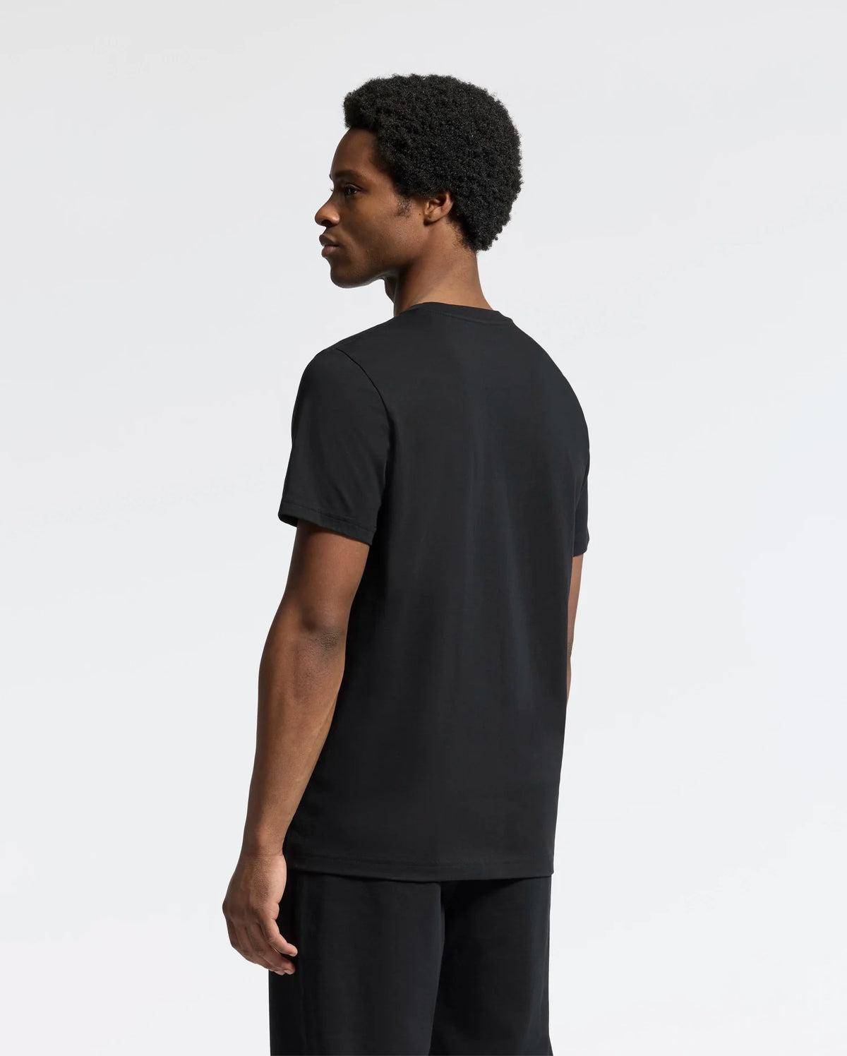 A person with short curly hair stands against a plain background, dressed in the Psycho Bunny MENS SYDNEY GRAPHIC TEE - B6U508F200 made of soft Pima cotton and dark pants, facing sideways to the left.
