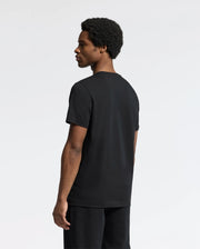 A person with short curly hair stands against a plain background, dressed in the Psycho Bunny MENS SYDNEY GRAPHIC TEE - B6U508F200 made of soft Pima cotton and dark pants, facing sideways to the left.