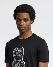 A person with short curly hair wears a Psycho Bunny MENS SYDNEY GRAPHIC TEE, a regular fit black Pima cotton T-shirt featuring a bunny with crossbones graphic. The background is plain and light-colored.