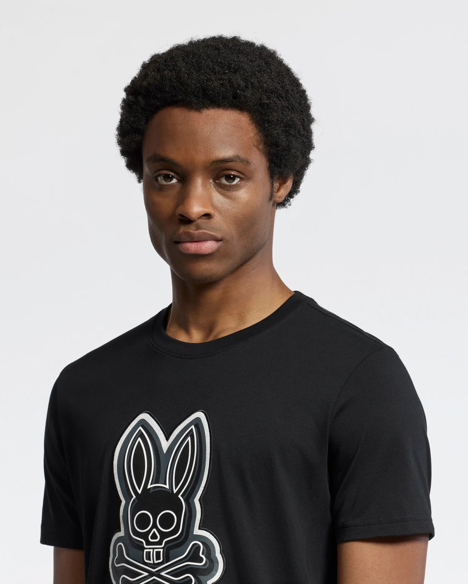 A person with short curly hair wears a Psycho Bunny MENS SYDNEY GRAPHIC TEE, a regular fit black Pima cotton T-shirt featuring a bunny with crossbones graphic. The background is plain and light-colored.