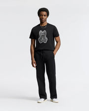 A person dressed in the Psycho Bunny MENS SYDNEY GRAPHIC TEE - B6U508F200, made of soft Pima cotton featuring a rabbit graphic, stands against a plain white backdrop. They're also wearing regular fit black pants and white sneakers.