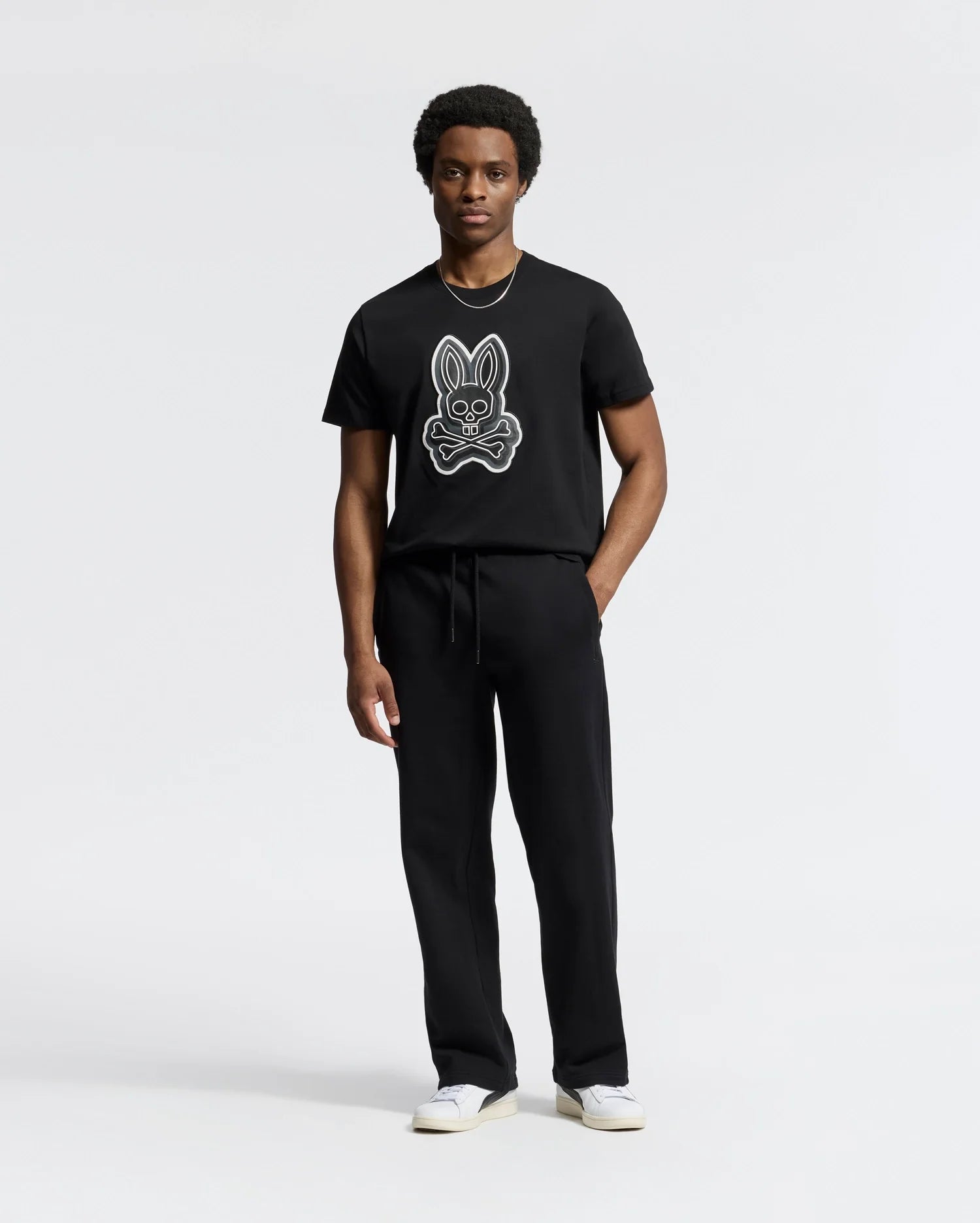 A person dressed in the Psycho Bunny MENS SYDNEY GRAPHIC TEE - B6U508F200, made of soft Pima cotton featuring a rabbit graphic, stands against a plain white backdrop. They're also wearing regular fit black pants and white sneakers.