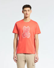 A young man wears the MENS SYDNEY GRAPHIC TEE - B6U508F200 from Psycho Bunny, a bold red Pima cotton shirt with a cartoon bunny and crossbones graphic on the chest. He pairs it with light beige pants, standing against a plain white background.