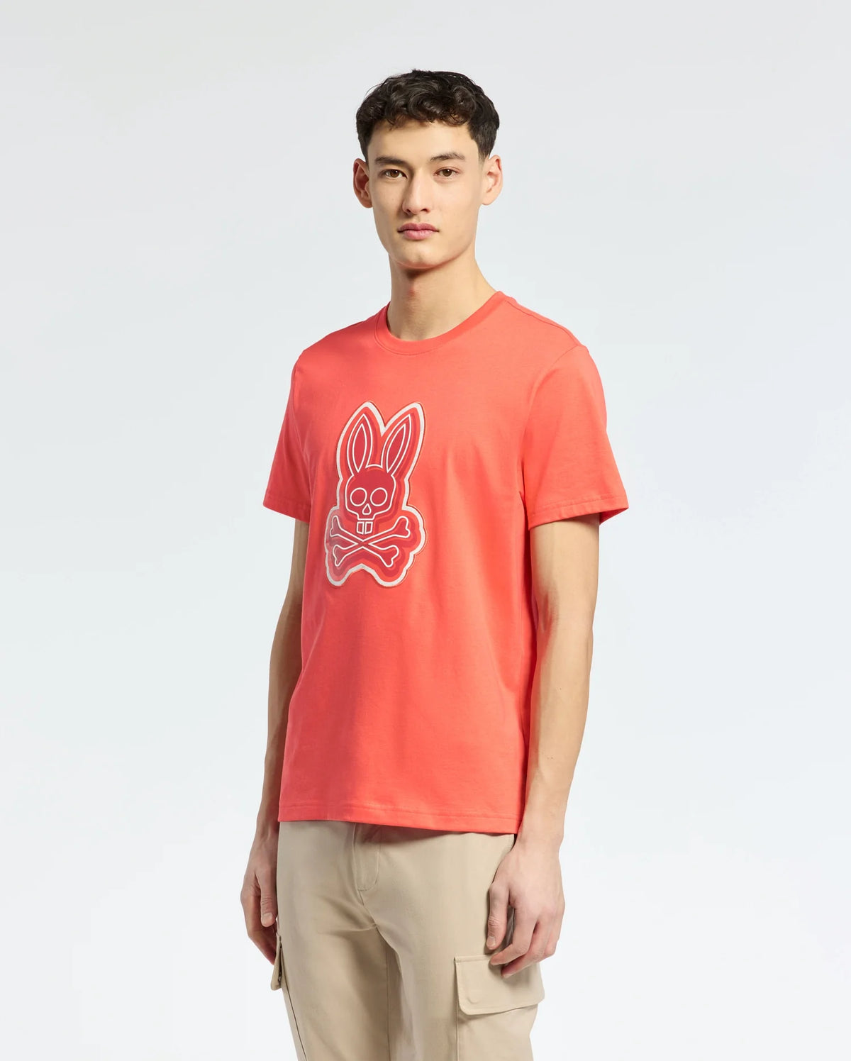 A person in a Psycho Bunny MENS SYDNEY GRAPHIC TEE - B6U508F200, orange with bunny and crossbones graphics, stands against a plain light background. Paired with beige cargo pants and short hair, they exude a casual style in this eye-catching ensemble.