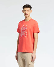 A person in a Psycho Bunny MENS SYDNEY GRAPHIC TEE - B6U508F200, orange with bunny and crossbones graphics, stands against a plain light background. Paired with beige cargo pants and short hair, they exude a casual style in this eye-catching ensemble.