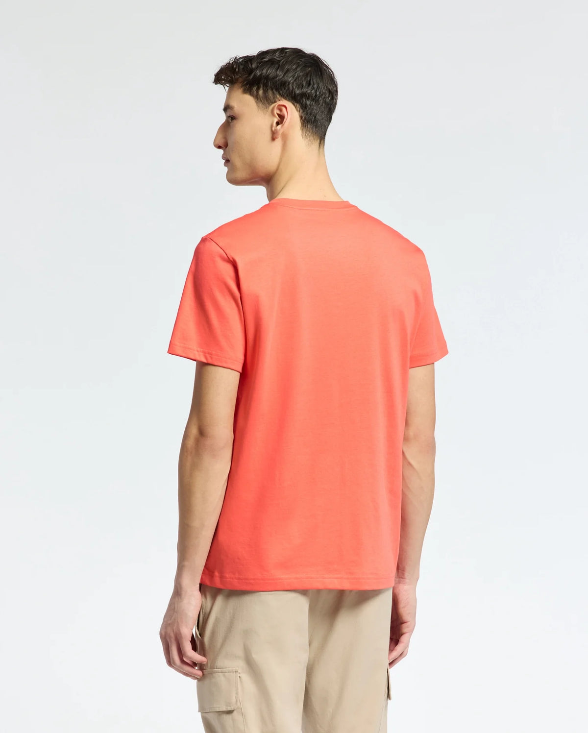A man faces away in the coral MENS SYDNEY GRAPHIC TEE by Psycho Bunny, made from soft Pima cotton, paired with beige cargo shorts. The plain white background contrasts his vibrant outfit.