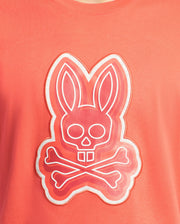 The Mens Sydney Graphic Tee by Psycho Bunny is a coral-colored T-shirt featuring a stylized graphic of a bunny with a skull face and crossbones. Made from soft Pima cotton, it displays layered pink shades with a white outline, offering an edgy twist on classic prints.