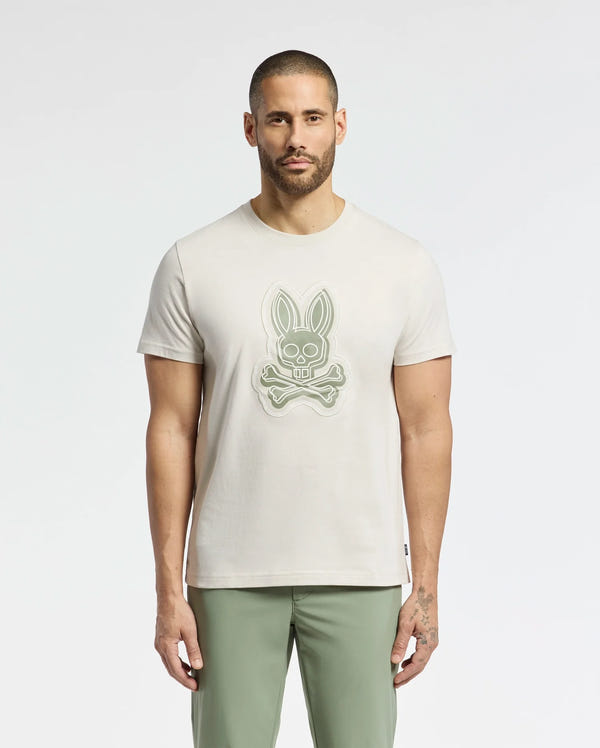 A man with short hair and a beard wears the Psycho Bunny MENS SYDNEY GRAPHIC TEE - B6U508F200 in beige, made from soft Pima cotton and featuring a bunny skull design. He pairs it with olive green pants against a light background.