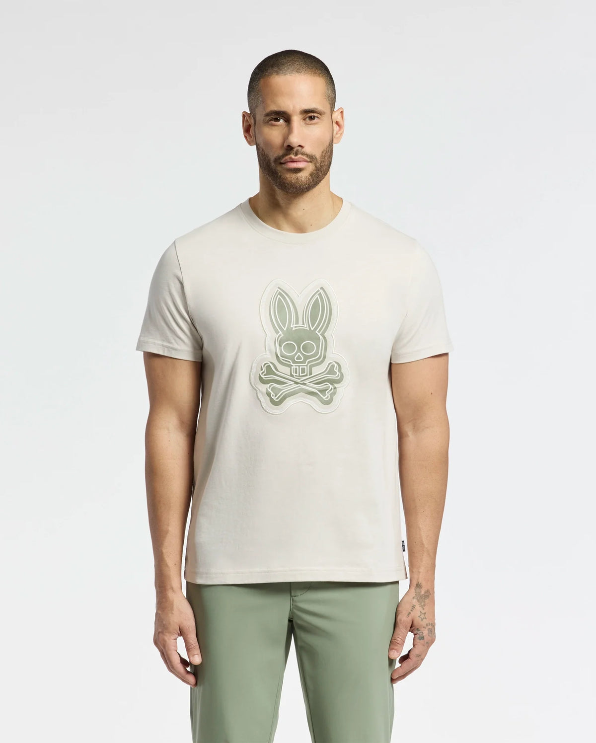 A man with short hair and a beard wears the Psycho Bunny MENS SYDNEY GRAPHIC TEE - B6U508F200 in beige, made from soft Pima cotton and featuring a bunny skull design. He pairs it with olive green pants against a light background.