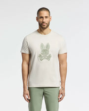 A man with short hair and a beard wears the Psycho Bunny MENS SYDNEY GRAPHIC TEE - B6U508F200 in beige, made from soft Pima cotton and featuring a bunny skull design. He pairs it with olive green pants against a light background.